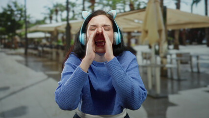 Woman wearing headphones shouting playfully on a terrace with blurred street and restaurant in the...