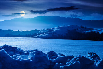 beautiful winter landscape with snow covered rolling hills under dark sky at night. majestic mountains in full moon light. frosty countryside scenery. backdrop for fiction art or mystery concepts