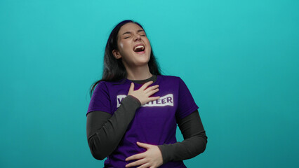 Obraz premium Woman singing passionately in purple volunteer shirt against blue background, expressing joy and emotion, brunette with long hair and hands to chest.