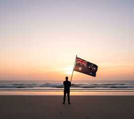 Lone figure stands silhouetted against a vibrant sunset holding a australian flag on a beach