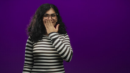 Young arab brunette woman wearing glasses and a striped top with hand covering mouth in studio; shyness.