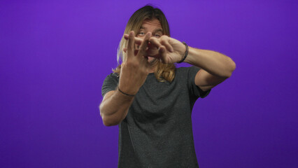 Man with long blond hair smiling and making hashtag gesture with fingers in purple studio; playful fun.