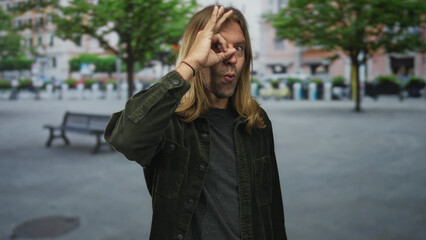 Fototapeta premium Blond haired man wearing green jacket makes ok sign with hand near eye on busy street; approval optimism.