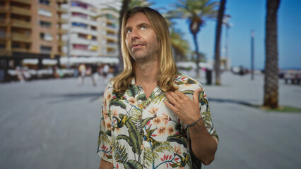 Fototapeta premium Man with long hair walking on promenade under bright sunlight by modern beachfront buildings; serenity.