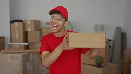 Man holding cardboard box and smiling in building wearing red cap and glasses amid stacked moving boxes; joy.