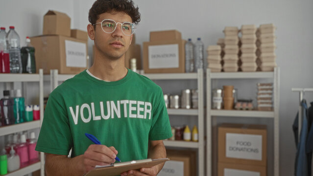 Young hispanic volunteer man wearing green shirt writes notes on clipboard in donation center room; compassion. - Powered by Adobe
