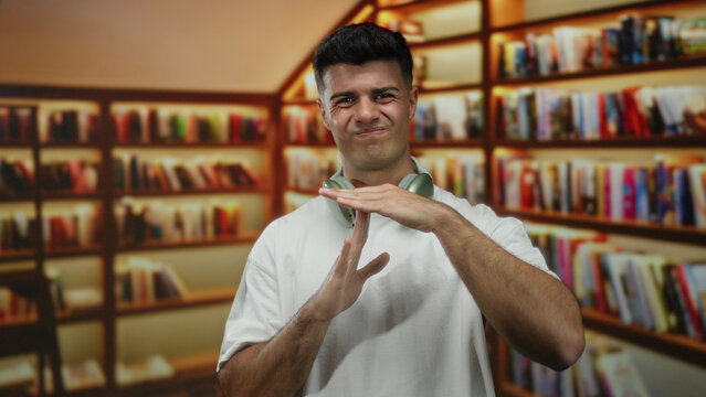 Young hispanic man gesturing time out with headphones in a library, surrounded by books, depicts a relaxed indoor academic setting with a handsome expression.