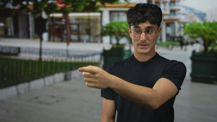 Young man hand on chin thinking gesture on street near planter and railing, wearing glasses and black t shirt; thoughtful reflection.