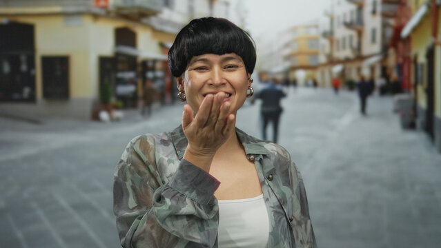 Woman blowing kiss on urban street with blurred cityscape background; young asian female depicted outdoors in vibrant city setting, suggesting lively urban lifestyle.