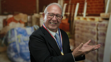 Naklejka premium Senior man in business suit proudly presents construction site indoors, wearing a medal, exuding confidence and leadership amidst building materials and bricks.