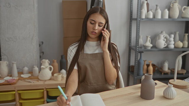 Young woman potter wearing apron holds phone to ear and writes with hand in notebook at a pottery studio surrounded by vases and shelves; contemplation planning. - Powered by Adobe
