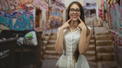 Naklejka premium Teen girl with braces points index fingers to her smile in a graffiti-lined street near stairs and a dumpster; playful confidence.