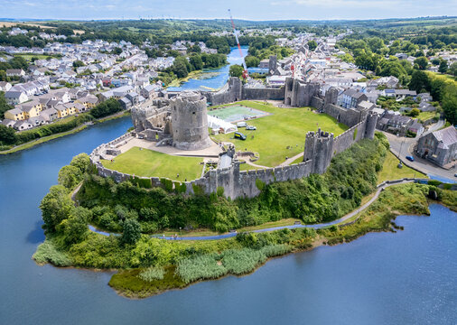 Aerial view of Pembroke Castle, its formidable stone walls embracing a verdant courtyard, reflected in the serene waters of the surrounding river, Pembroke, Pembrokeshire, Wales.
