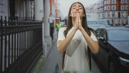 Fototapeta premium Woman with hands clasped praying on a street in a city near parked cars and an iron fence; reflection hope serene.