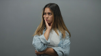 Woman standing with hand on cheek and folded arm wearing denim shirt in gray studio; pensive...