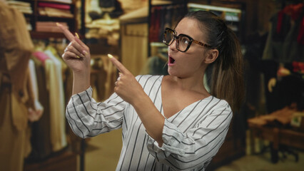 Young woman points finger across clothing racks in a retail building, wearing glasses and striped blouse, open mouth expression; surprise discovery.