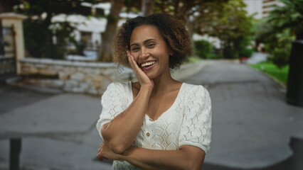 Woman smiling with hand on cheek on street near stone wall and trees, arms folded and laughing on...