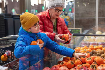 Grandmother and Grandson Selecting Persimmons Together in the Supermarket.  Shopping. Family Support, Healthy Eating, Active Senior Lifestyle, and Intergenerational Connection Between Gen Alpha 
