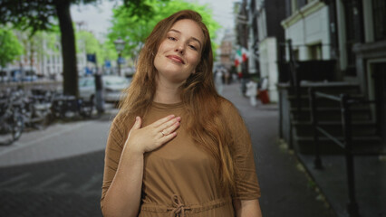 Fototapeta premium Smiling woman places hand on chest wearing brown dress on quiet city street in early morning light; gratitude.