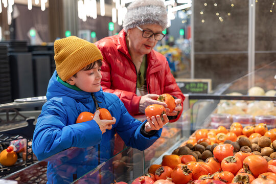 Grandson Helping His Grandmother Choose Fresh Persimmons in a Supermarket Produce Section &ndash; Family Support, Healthy Eating, Active Senior Lifestyle, and Intergenerational Connection Between Gen Alpha 