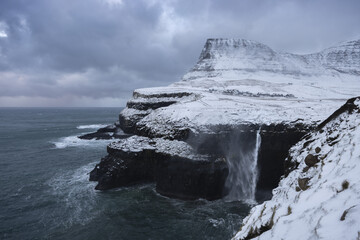 View of a dramatic waterfall cascading into the sea from snow-covered cliffs under a cloudy sky, a winter scene of stark beauty, Sorvagur, Vagar, Faroe Islands.