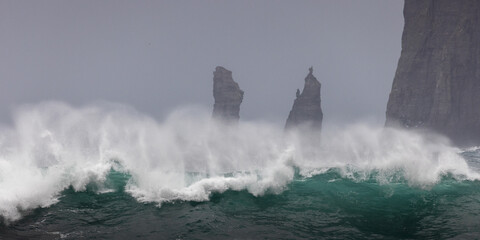 View of powerful turquoise waves crashing against rugged cliffs and sea stacks under a misty sky, creating a dramatic coastal scene, Sorvagur, Vagar, Faroe Islands.