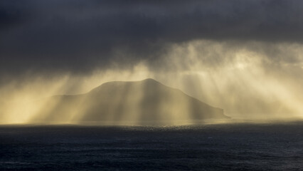 View of sunbeams piercing through the dark, stormy clouds over the sea, illuminating a distant island in a dramatic display of light and shadow, Sorvagur, Vagar, Faroe Islands.