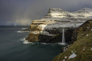 View of stark cliffs meet the churning sea, a waterfall plunges from snow-dusted heights under a brooding sky, hints of a rainbow paint the scene, Sorvagur, Vagar, Faroe Islands.