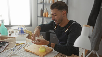 Young hispanic man shaping white clay with bare hands on wooden table in workshop; creative craftsmanship focus.
