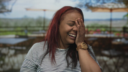 Fototapeta premium African american woman covers mouth laughing on restaurant terrace building outdoors; lighthearted joy.