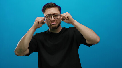 Naklejka premium Young man with beard making crying gesture against a blue background, expressing emotion with hands near face, wearing casual black shirt.