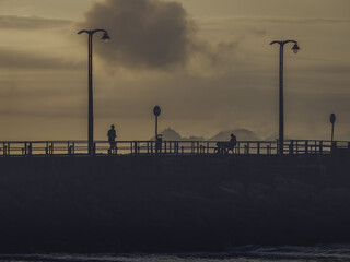 View of figures silhouetted against the muted golden light of dusk on a pier, the calming ocean waves reflecting the sky, Illa de Toralla, Pontevedra, Spain.