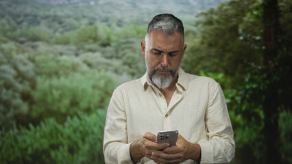 Fototapeta premium Man in linen shirt looking down at smartphone and holding it with both hands in a forest setting among green bushes and grassy field; concentration.