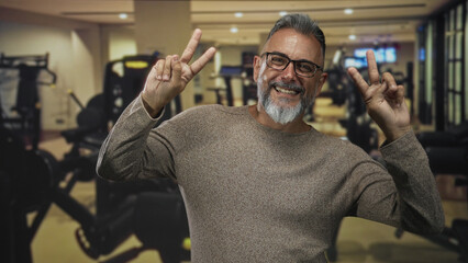 Man middle aged with beard and glasses flashing peace sign with both hands while smiling in gym building; joy wellness.
