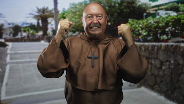 Elderly man monk in brown robe raises clenched fists and displays a large wooden cross necklace along a stone walled street lined with palm trees; triumph faith.