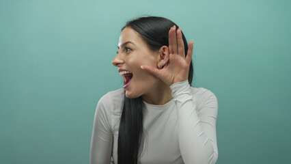 Young hispanic woman wearing white shirt smiles while holding hand to ear in a playful listening gesture against an isolated green background wall.