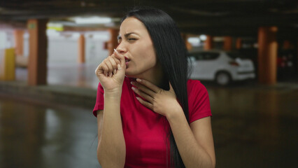 Fototapeta premium Woman coughing in an indoor parking lot beside a parked car, wearing a red shirt and appearing distressed, set against an urban background, highlighting human emotion and health.