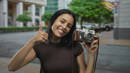 Young woman holding camera smiling with thumbs up on city street during daytime showcasing urban...