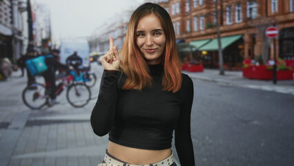 Redhead woman smiling points finger upward on a busy urban street beside a cyclist and a parked car, facing camera in a cropped midshot; confidence.