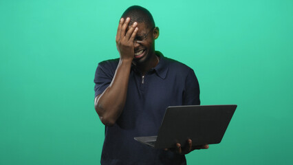 Man holding laptop with hand covering face and glasses, facepalm gesture in studio; frustration overload.