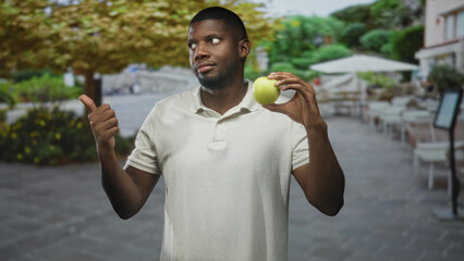 Black man holding a green apple and showing thumb up on street cafe patio wearing white polo while facing camera; confidence approval.