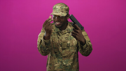 Fototapeta premium Man soldier in camouflage uniform holding a gun to his head while making a pinching fingers gesture against a magenta studio backdrop, smiling; tension threat.