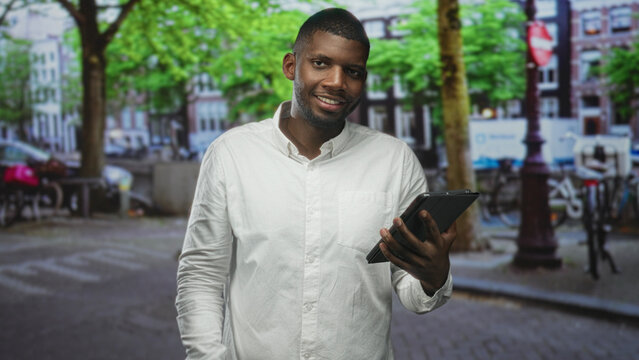 Man tapping tablet with finger on street with parked bicycles and trees, wearing white shirt and smiling; connectivity concentration. - Powered by Adobe