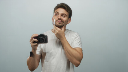 Man holding camera and touching chin in studio with pale blue backdrop wearing white t shirt; thoughtful reflection.
