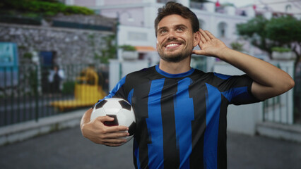 Young hispanic man wearing blue and black soccer jersey holding a soccer ball and smiling on street by building fence and rail; team spirit joy.