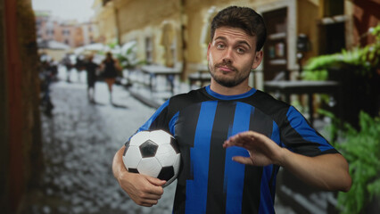 Man holding soccer ball with palm out for stop on a cobbled city street in blue striped jersey; refusal defiance.