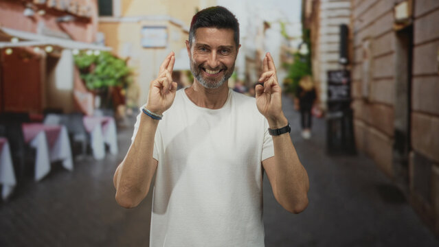 Man shows crossed fingers with both hands while smiling on street outside restaurant terrace; hopeful anticipation.