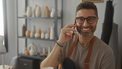 Man holding smartphone to ear, smiling in studio with pottery on shelves while wearing apron and glasses; creative connection joy.