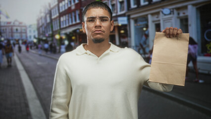 Young hispanic man holds paper bag aloft with right hand on a busy street, wearing glasses and cream sweater, inspecting the bag; urban confidence.