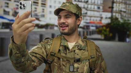 Fototapeta premium Man in military uniform holds smartphone for a video call on a busy urban street lined with apartment buildings; connection communication camaraderie service.